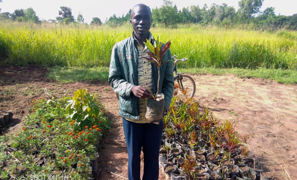 young man holding a seedling plant standing out in the open with lush green setting in the background