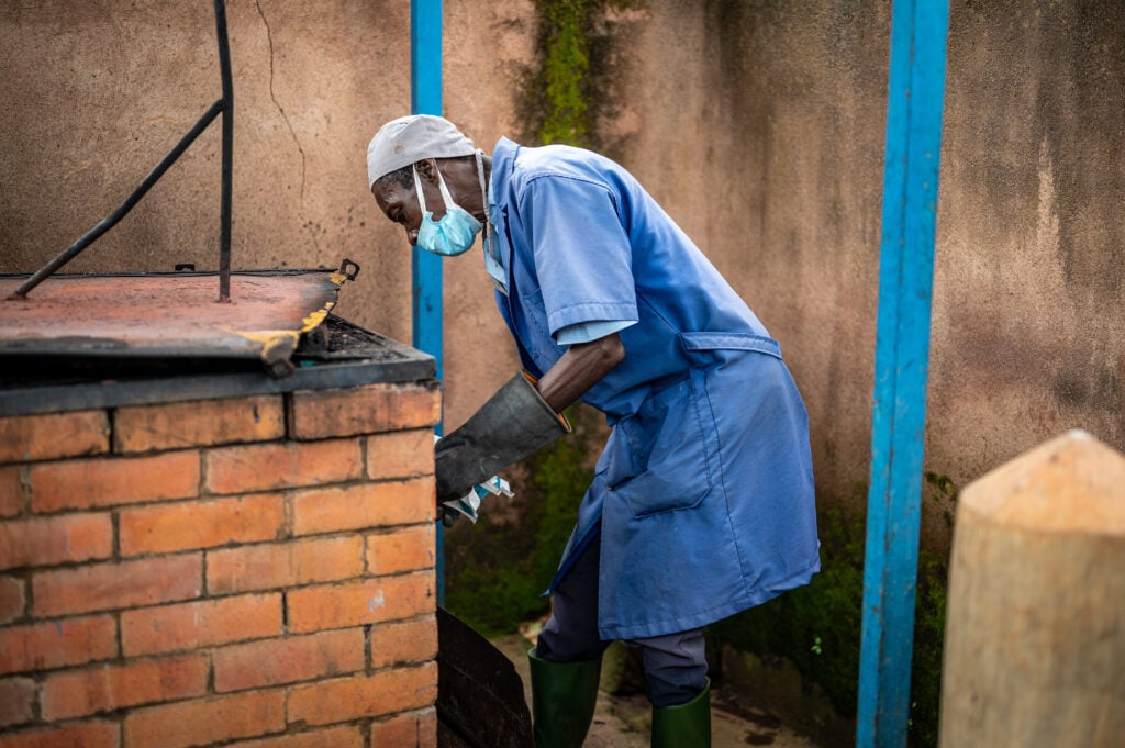 man with face mask and gloves cleans what looks like an incinerator in acompound