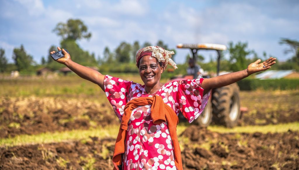 smiling woman standing in a farm plot holding a mobile and spreading both arms into the air