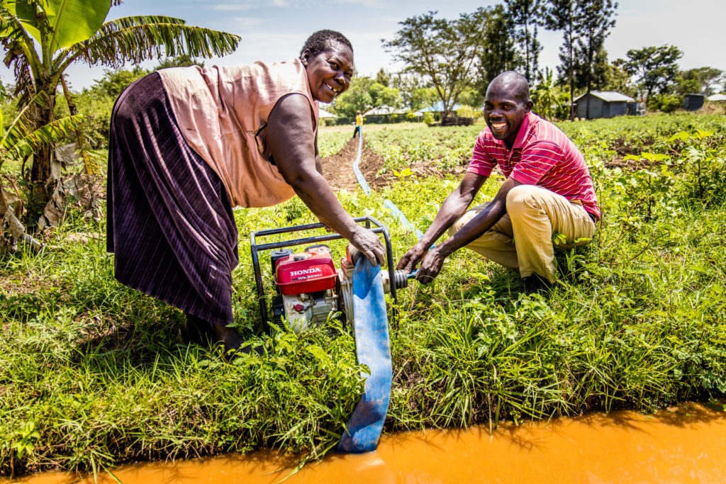 a woman and a man in a farm field standing close to an irrigation channel with a water pump