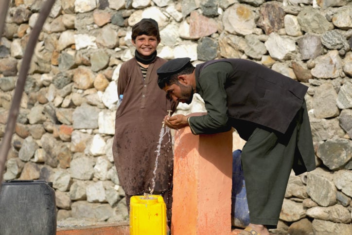 man stooping to drink from a tap and a young boy standing behind him smiling