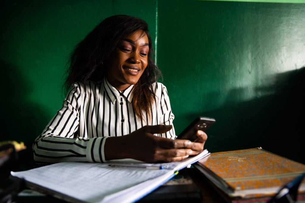 young woman behind desk with notebooks is smiling and watching her phone