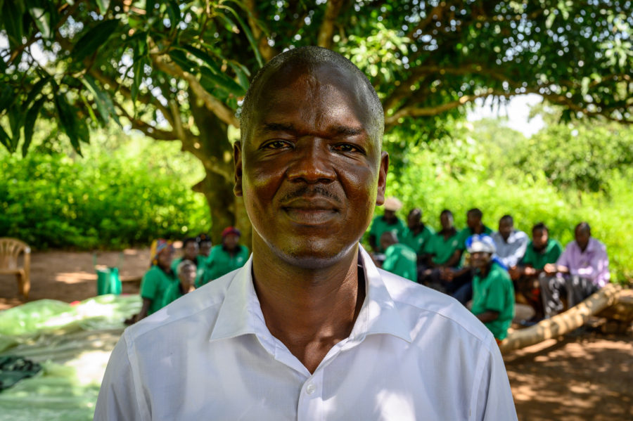 man looking into the camera smiling with a group of people in the background in the shade under a tree