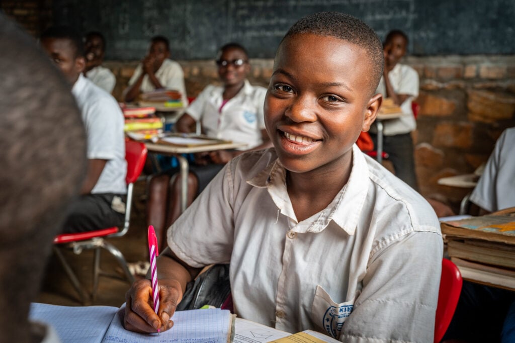 en lachend meisje in een wit schooluniform zit in een klaslokaal, met een roze pen in de hand en een open schrift op tafel. Achter haar zijn andere leerlingen zichtbaar die aan hun schoolwerk zitten. Het lokaal heeft stenen muren en een schoolbord.