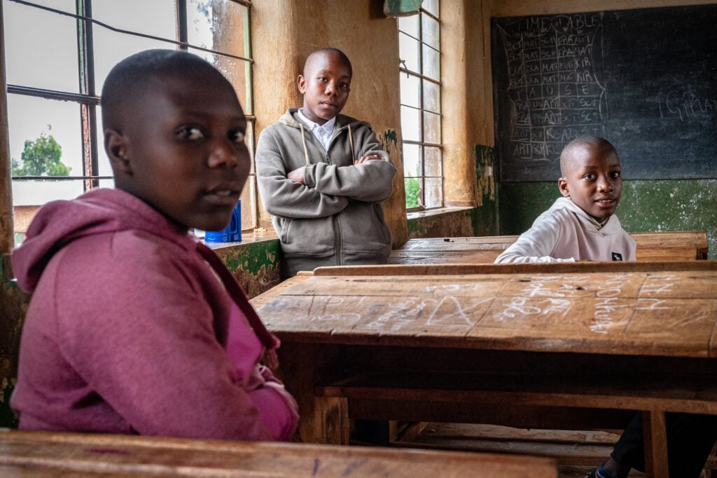 Drie kinderen zitten in een klaslokaal met houten banken. Een jongen staat leunend tegen een raam met gekruiste armen, terwijl de andere twee kinderen zitten en naar de camera kijken. Het lokaal heeft een verweerde muur, schoolbord en grote ramen.