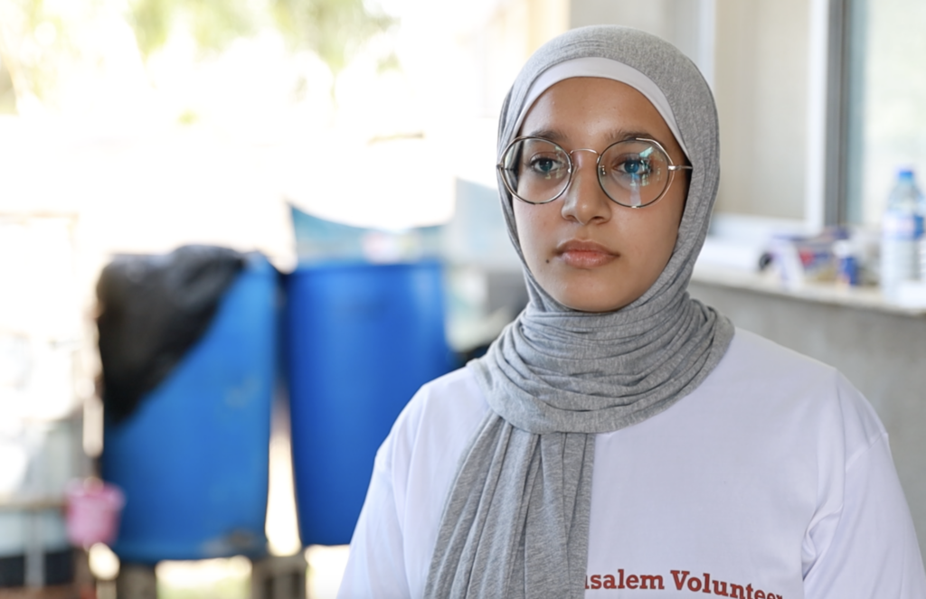 Een jonge vrouw met een grijze hoofddoek en bril. Ze heeft een wit T-shirt aan met de tekst "Jerusalem Volunteers". En zit in een lichte ruimte. Op de achtergrond zijn blauwe tonnen en onscherpe details zichtbaar.