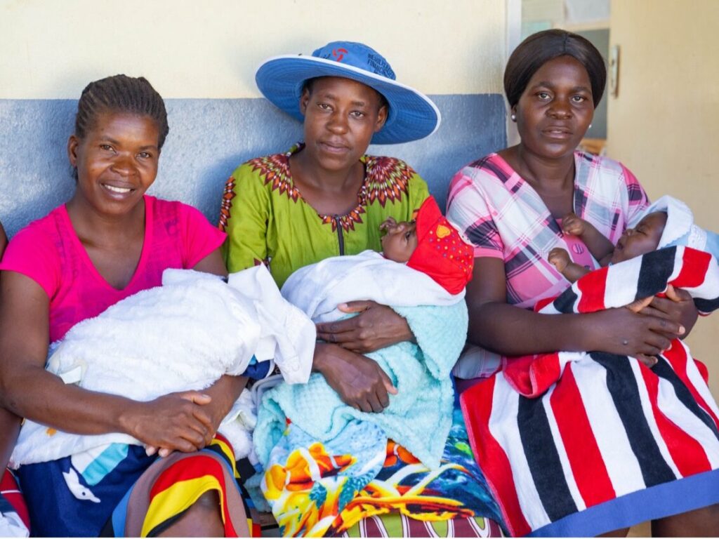 Drie moeders in Zimbabwe zitten naast elkaar, elk met hun baby op schoot, lachend en trots, als resultaat van verbeterde toegang tot gezondheidszorg via het Health Sector Development Project.