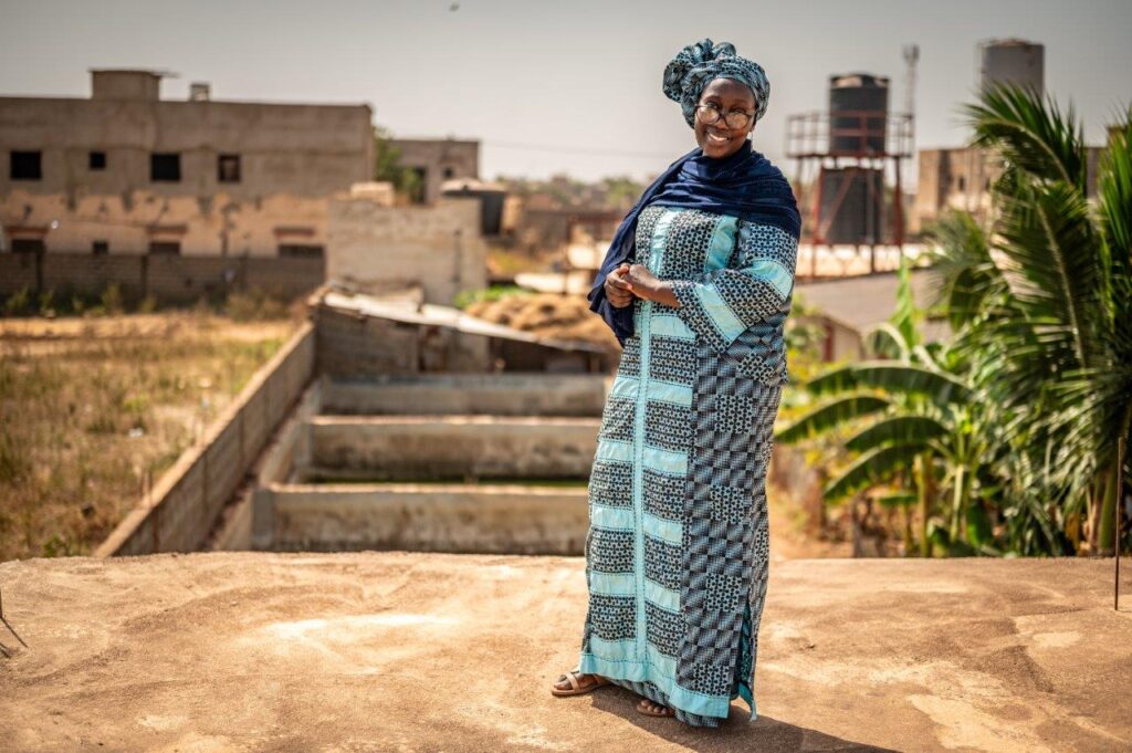 Een vrouw glimlacht terwijl ze buiten poseert. Ze draagt een kleurrijke traditionele outfit in blauwe tinten met een bijpassende hoofddoek. Op de achtergrond is een stedelijk landschap te zien met gebouwen, een muur en weelderige groene planten zoals palmbomen. De sfeer is zonnig en warm.