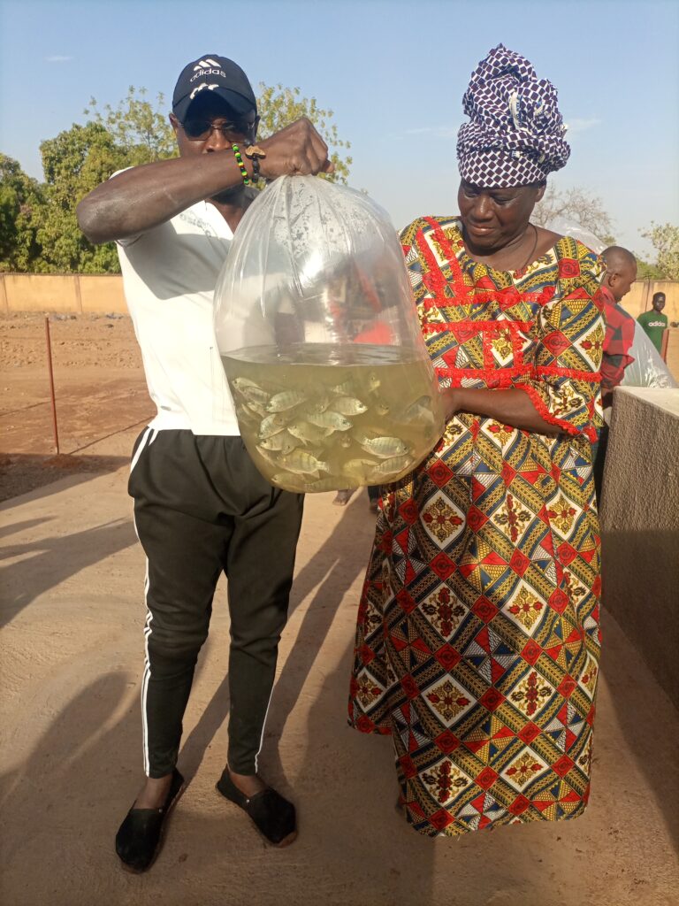 Een man en een vrouw uit Burkina Faso houden samen een grote, doorzichtige plastic zak vast die gevuld is met water en levende vissen. De vrouw draagt een kleurrijke jurk met een bijpassende hoofddoek, de man een pet en sportieve kleding. Ze staan buiten op een zonnige dag in een droge omgeving met enkele bomen op de achtergrond.